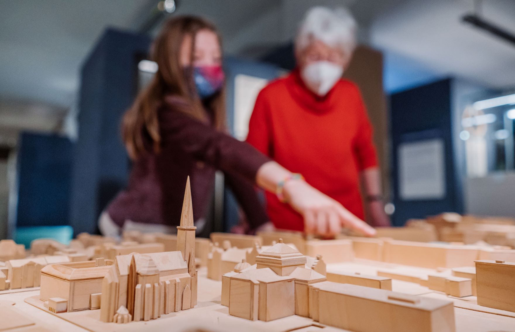 Im Vordergrund das Modell einer Stadt aus Holz, im Hintergrund eine Oma mit Enkelin