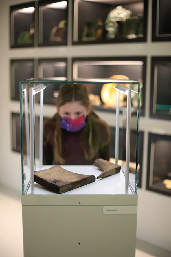 Ein Kind guckt durch eine Vitrine, in der eine alte Klobrille aus einer Latrine liegt.