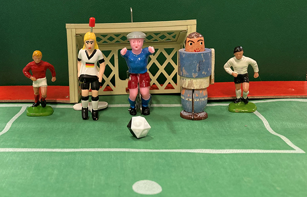 5 sehr unterschiedlich aussehende Spielfiguren stehen vor dem Tor eines Tischfußballspieles.