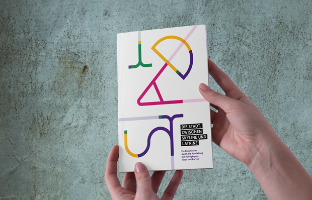 H&auml;nde halten den Katalog zur Ausstellung: ein wei&szlig;es Buch mit bunten Buchstaben, die das Wort STADT bilden