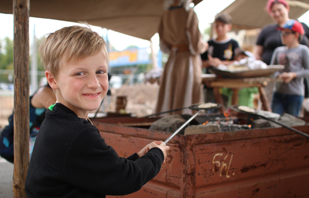 Das Bild zeigt einen grinsenden Jungen, der einen Brotfladen über einem Kohlefeuer bäckt. Foto: Annelie Blasko
