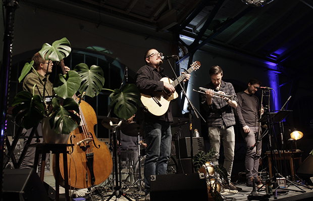 Das Foto zeigt den Auftritt einer Band. Zu sehen sind ein Mann an der Gitarre, der in ein Mikrofon sing. Zwei weitere M&auml;nner halten Blasinstrumente in der Hand, spielen aber gerade nicht.