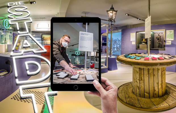Foto einer Hand, die ein Tablet h&auml;lt. Im Hintergrund ist die Ausstellung zu sehen.