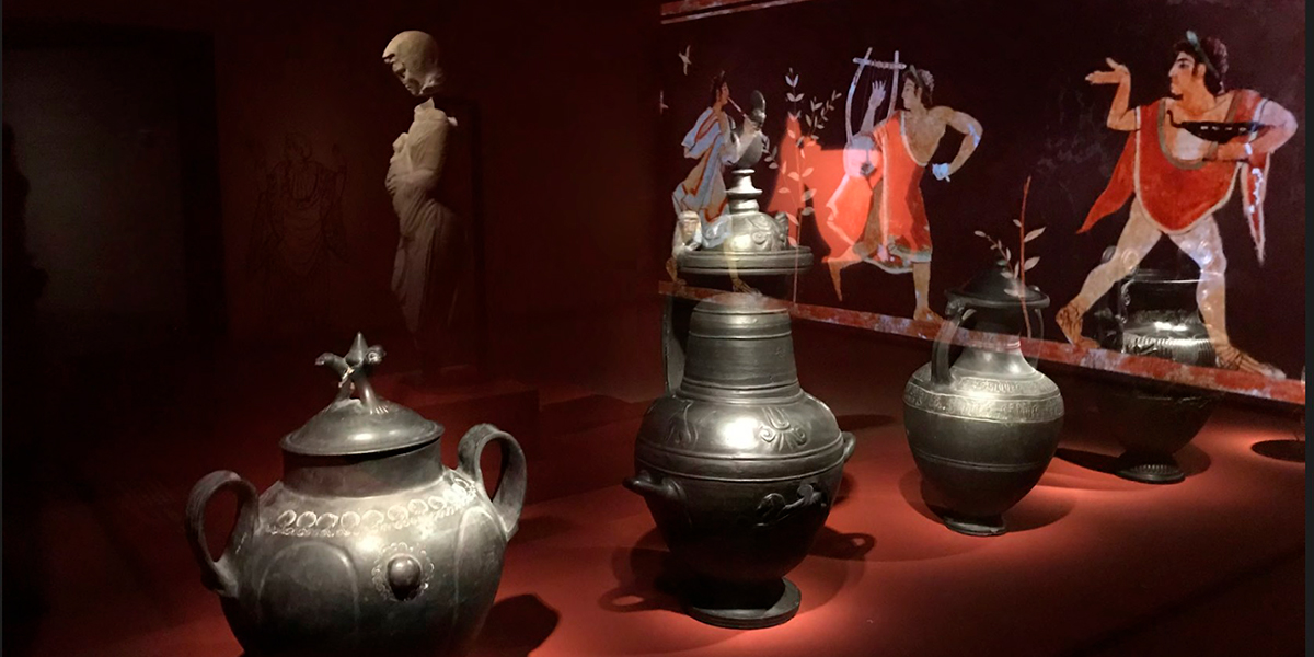Im Vordergrund stehen antike Bronzegef&auml;&szlig;e, im Hintergrund zeigt eine Wandmalerei mehrere musizierende Personen. Eine Statue ist weiter hinten zu erkennen.