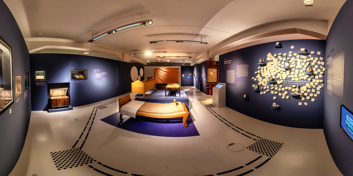 Das Foto zeigt einen Raum in der Ausstellung "Home Sweet Home". Seine Wandelemente sind dunkelblau. Mehrere ungewöhnlich aussehende Betten sind zu erkennen.