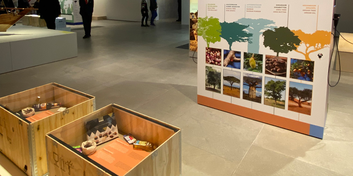 Blick auf den Boden eines Ausstellungsraumes. Hier stehen 2 Installationen: Holzkisten mit afrikanischen Produkten darin. Außerdem eine Textstele mit Bildern von afrikanischen Baumarten.