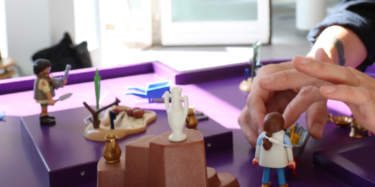 Auf einem lila-farbenen Untergrund deponiert eine Hand Playmobil-Figuren.