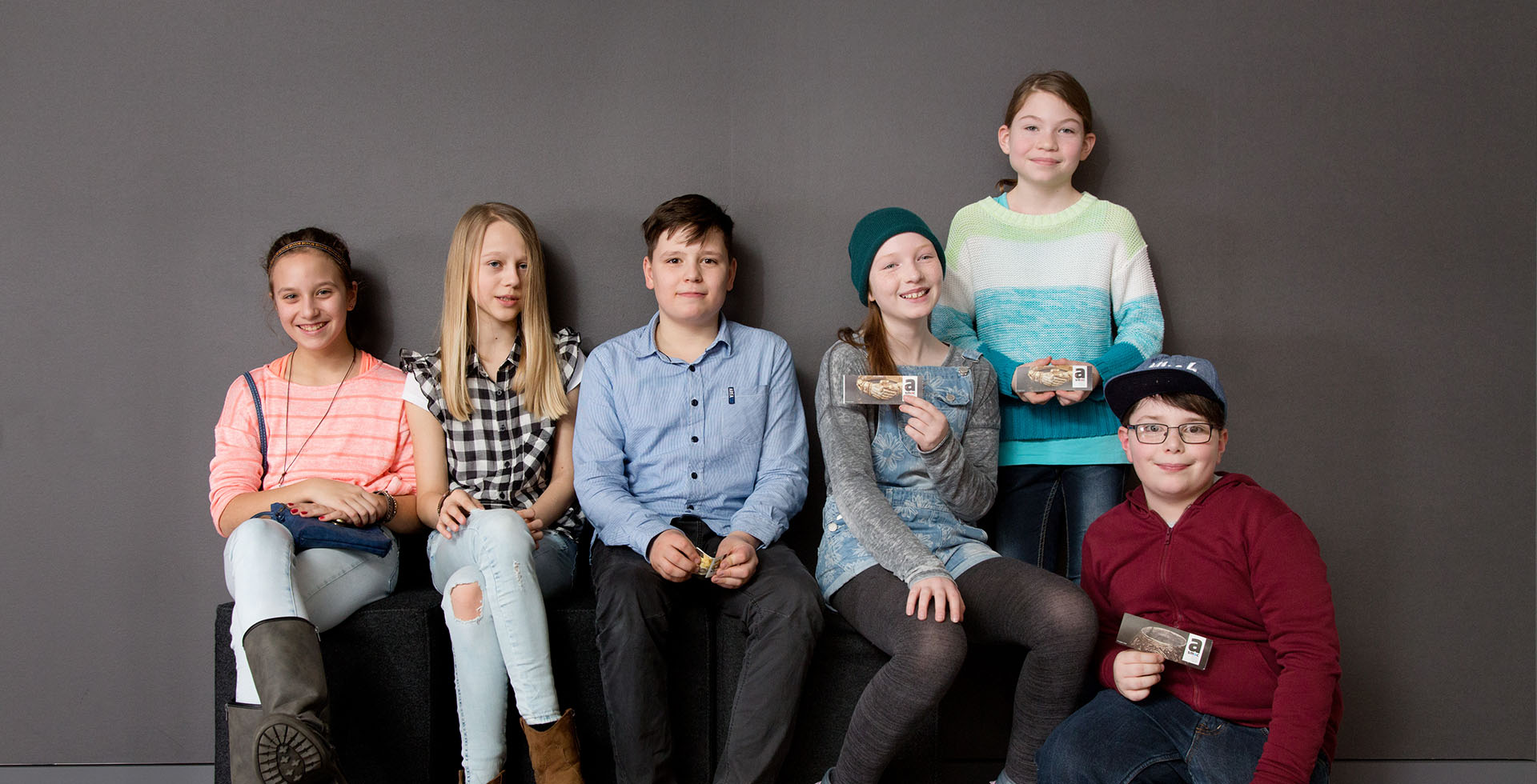 Sechs 10-jährige Kinder sitzen, stehen und hocken vor einer grauen Wand. 3 von ihnen halten eine Eintrittskarte in der Hand.