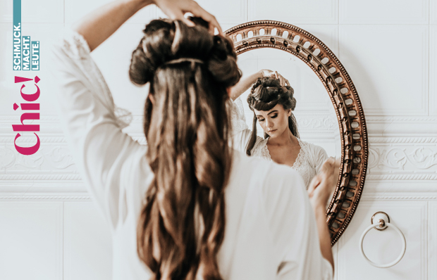 Eine junge Frau in weißem Kleid steht vor einem Spiegel und richtet sich die Haare.