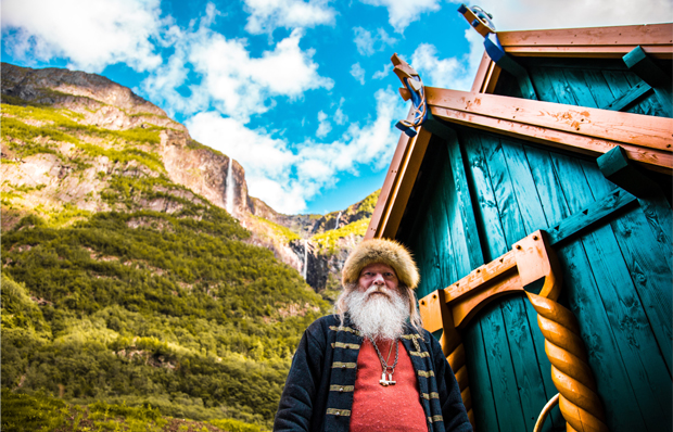 Ein alter Mann mit weißem Rauschebart und Pelzkappe steht vor einem altertümlichen türkisen Holzhaus. Das Haus steht in einer felsigen Landschaft.