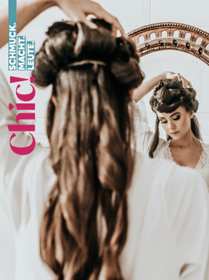 Eine junge Frau in weißem Kleid steht vor einem Spiegel und richtet sich die Haare.