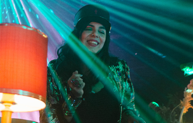 Das Foto zeigt eine Situation in einem Club: eine junge Frau mit langen dunklen Haaren und Käppi steht hinter den Plattentellern. Ein Scheinwerfer wirft Lichtstrahlen auf sie. Sie genießt ihre Musik.