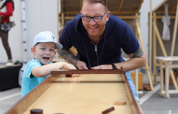 Ein Vater und sein Kind - ein Junge mit weißer Baseballkappe - spielen an einem langen Spieltisch. Man muss runde Spielsteine durch Tore schubsen. Beide sind fröhlich.