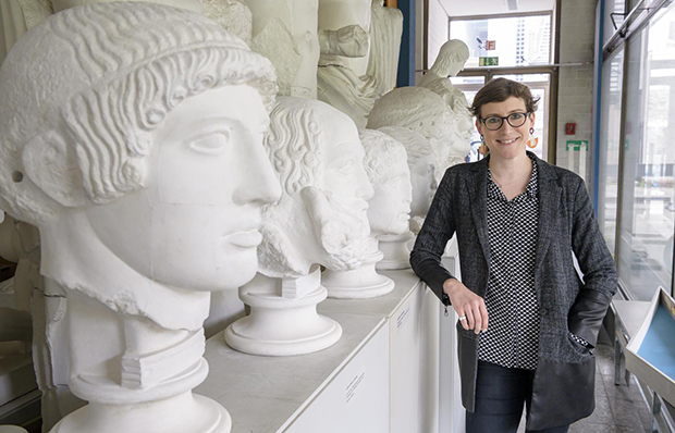 Eine junge Frau mit kurzen, dunklen Haaren und Brille steht neben einem Regal mit Gipsabgüssen von Köpfen großer römischer Statuen.