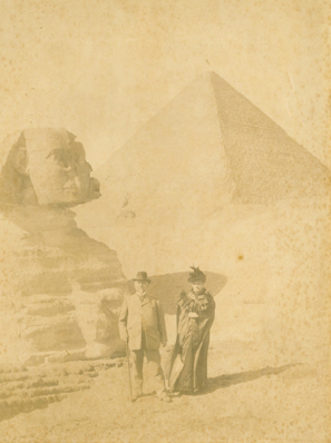 Alte vergilbte Fotografie. Ein Mann und eine Frau stehen mit Kleidung im Stil um 1900 stehen vor dem Sphinx in Gizeh, Ägypten. Im Hintergrund ist eine Pyramide zu sehen.