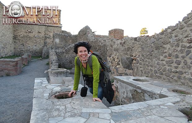 Eine mittelalte Frau mit kurzen, dunklen Locken stützt sich mit den Händen auf eine Mauer und lacht in die Kamera. Sie steht inmitten der Ruinen von Pompeji.
