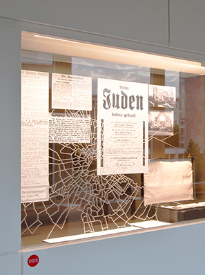 In einer großen Wandvitrine hängen Schriftdokumente, unter anderem eines mit der Überschrift „Beim Juden haben gekauft…“ mit einer Namenslist. Auf der Vitrinenscheibe klebt ein spinnennetzartiges Geflecht, das einen Sprung im Glas nachahmt.