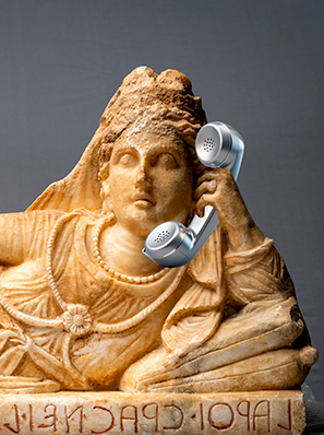 Liegende weibliche Statue aus Alabaster, die sich auf ihren linken Ellbogen stützt. Sie trägt ein faltenreiches Gewand, viel Schmuck und einen Schleier. Sie hält einen Telefonhörer in der Hand.