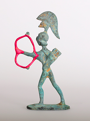 Die bronzene Statuette eines Kriegers trägt einen Helm mit hohem Kamm und auf dem Rücken einen Köcher. Der Bogen, den er eigentlich spannt, wurde durch ein rotes Herz ersetzt.